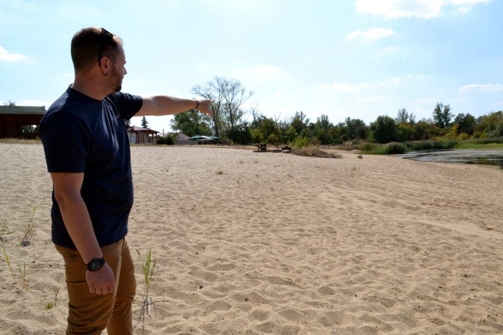 Fabian Schauer zeigt auf einen Stein unter einem Busch, der weit entfernt vom Wasser im Sand liegt und die frühere Wassergrenze markiert.