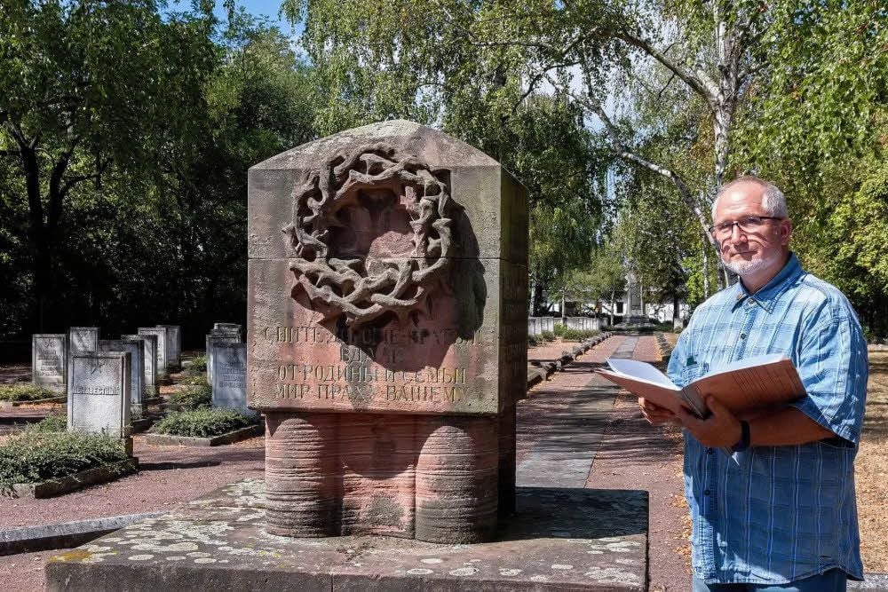 Dietmar Eißner auf dem Ehrenfriedhof: Neben ihm ein Gedenkstein, den russische Lagerinsassen, die nach Kriegsende 1918 nicht gleich nach Hause konnten, im Gedenken an ihre toten Kameraden hatten aufstellen lassen. In Eißners Rücken liegt das Gräberfeld mit den verstorbenen russischen Lagerinsassen aus dem Ersten Weltkrieg. Weltkriegstote anderer Nationalitäten liegen nicht mehr hier.