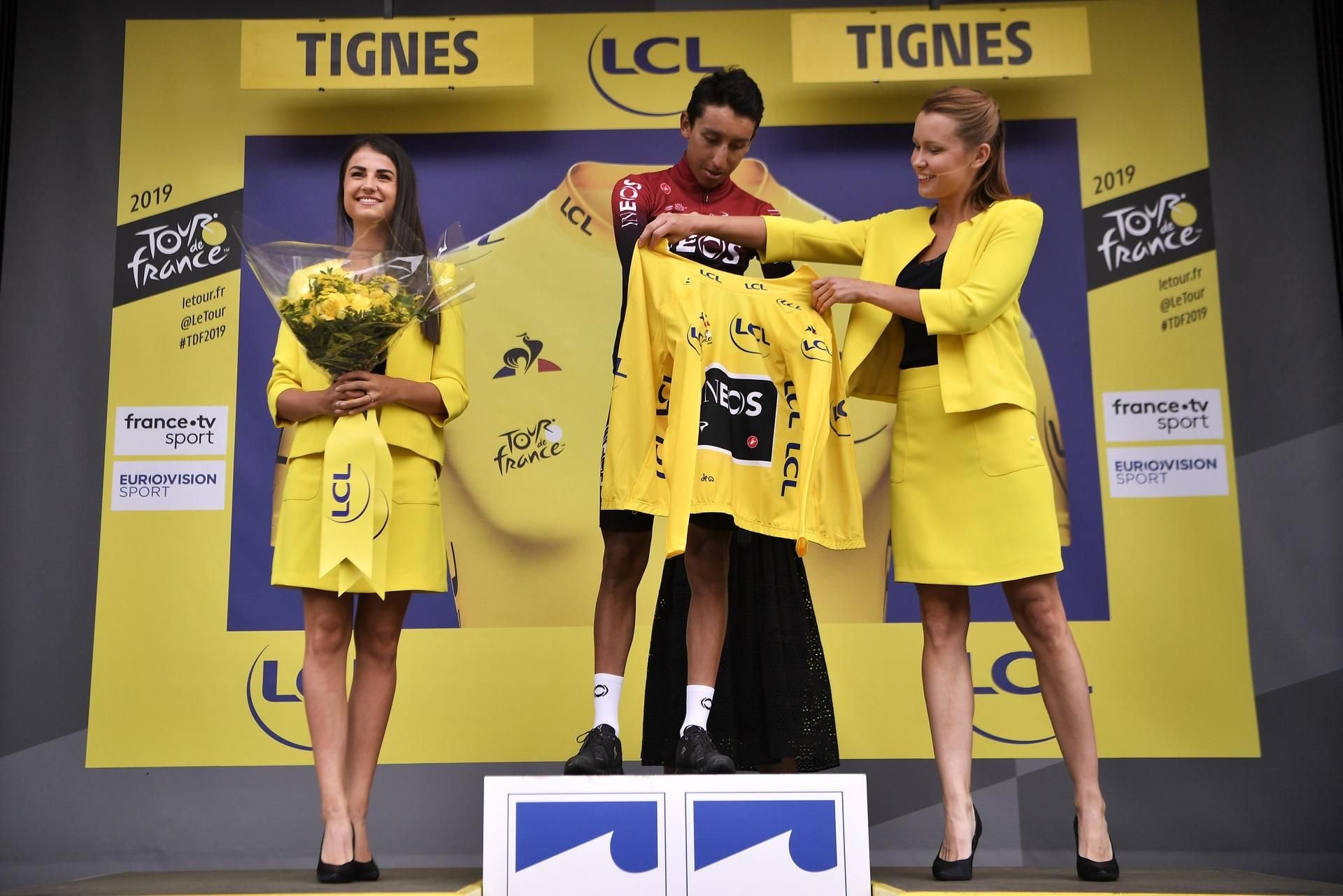 Egan Bernal (M.) bekommt im Juli 2019 sin Gelbes Trikot von zwei Hostessen überreicht.