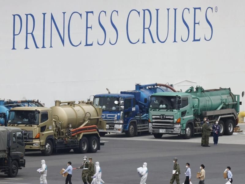 Menschen mit Mundschutz und teilweise mit Schutzanzügen gehen im Hafen von Yokohama an dem unter Quarantäne stehenden Kreuzfahrtschiff „Diamond Princess” vorbei. Foto: ---/kyodo/dpa