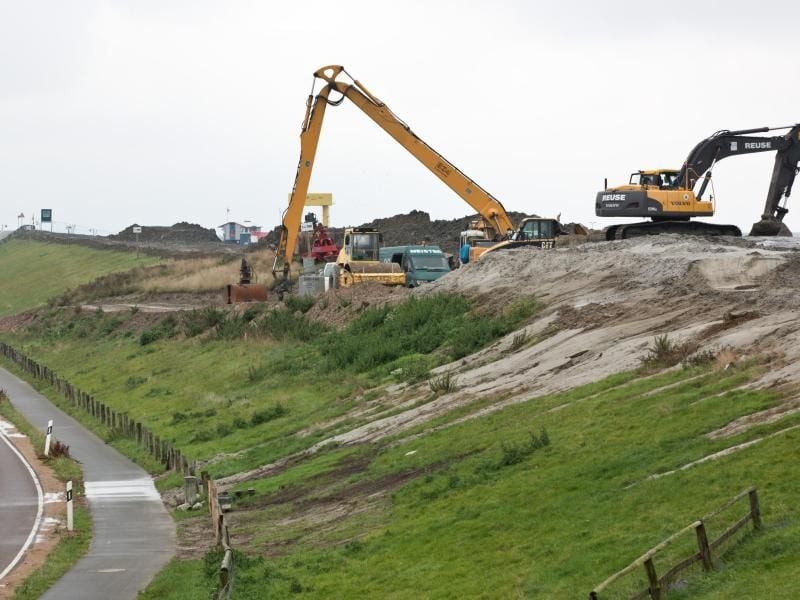 Auf einer Deichbaustelle tragen Arbeiter mit schwerem Gerät die Deichkrone des alten Deiches ab. Foto: Christian Charisius/Archiv