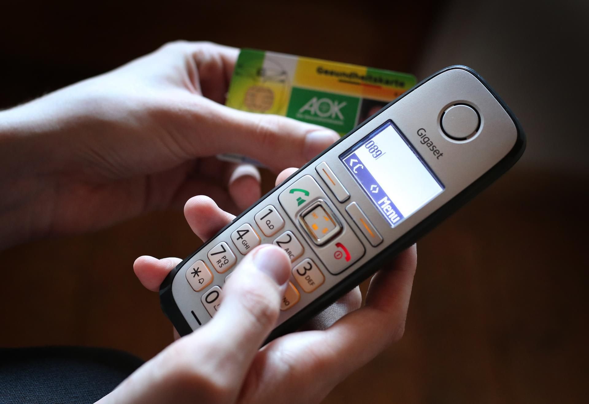 Illustration zum Thema telefonische Krankschreibung: Ein Mann hält ein Telefon und eine Gesundheitskarte der AOK in der Hand.