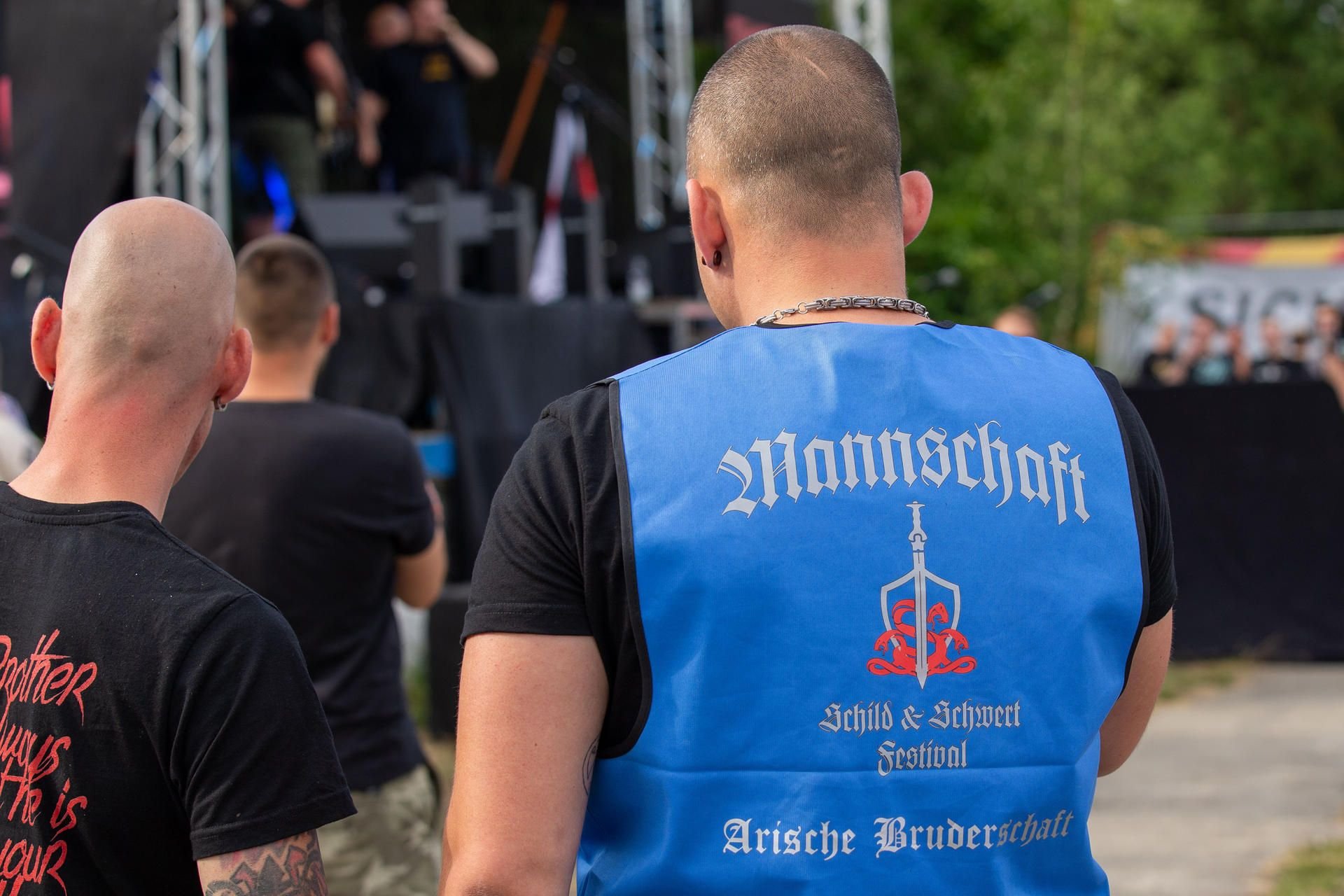 Auf der Weste des rechten Mannes steht auf dem Rücken die Aufschrift: Mannschaft - Schild und Schwert Festival - Arische Bruderschaft.