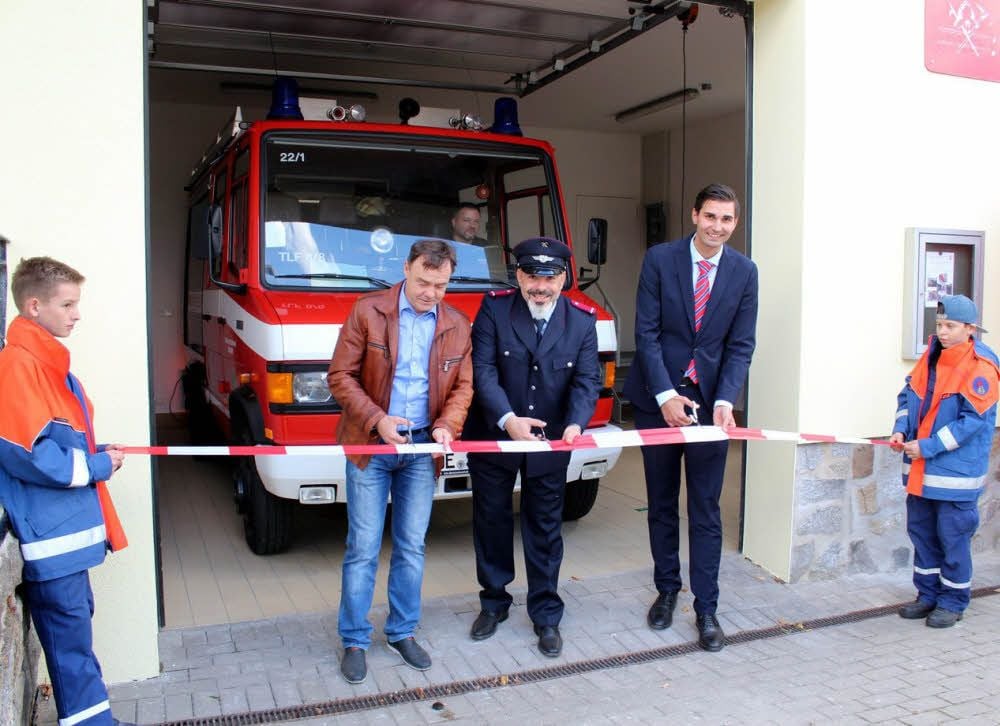 Bürgermeister Michael Jahn, Ortswehrleiter Oliver Asmus und Bundestagsabgeordneter Sepp Müller zerschneiden das Band. Kurz darauf fährt das neue Prunkstück aus seiner tollen Garage.