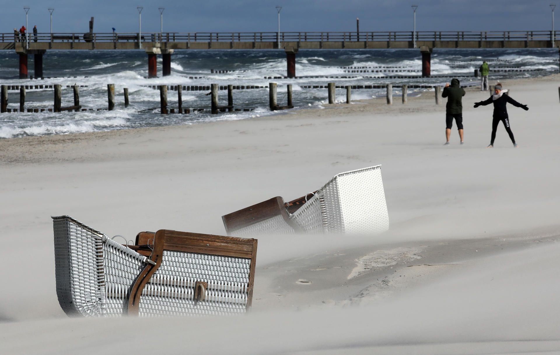Umgefallene Strandkörbe liegen im Sand, während Spaziergänger bei Sturm und Regen am Ostseestrand spazieren gehen.
