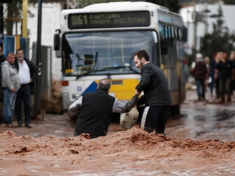 Ein Mann hilft an einer überflutete Straße in Mandra einem anderen Mann. Foto: Eurokinissi/Zuma