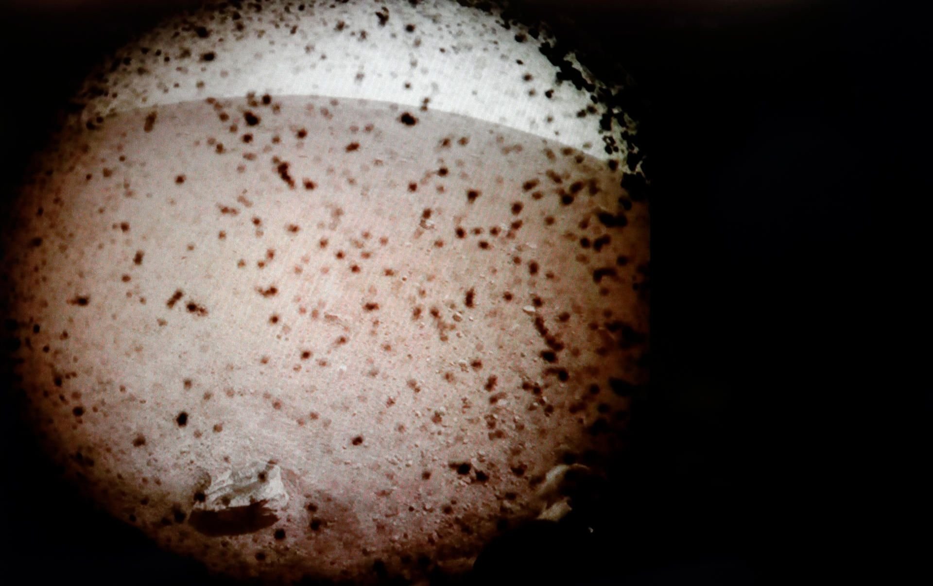 Das erste Bild, das vom InSight Mars-Lander aufgenommen wurde, nachdem er auf der Oberfläche des Mars landete. Trümmer, die von den Raketen des Landers aufgewirbelt werden, bedecken den Schutzschild der Kamera, der später entfernt wird.