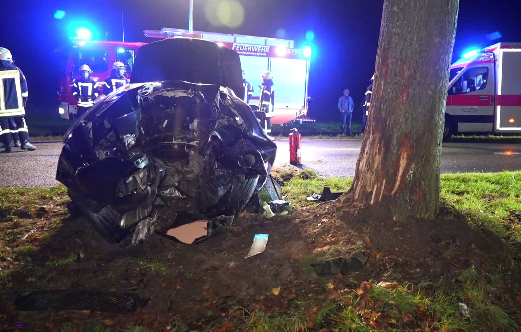 Rettungskräfte stehen an der Unfallstelle in Syke neben dem völlig zerstörten Unfallauto.