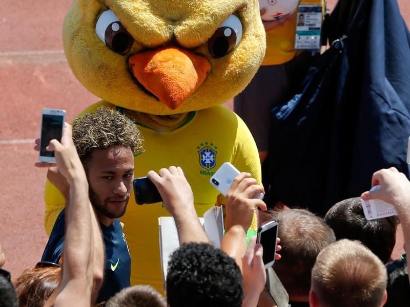 „Flügelmann” Canarinho, das Maskokttchen der brasilianischen Nationalmannschaft, ein etwas böse blickender Kanarienvogel, wacht über Fußball-Superstar Neymar, der beim Trainingsgeländer von Fans belagert wird. Foto: Andre Penner