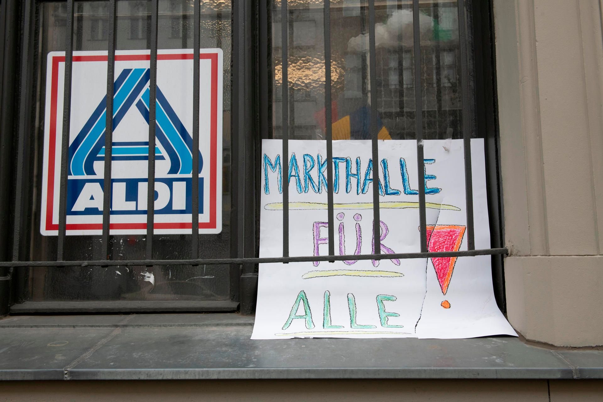 Ein selbstgemaltes Schild mit der Aufschrift «Markthalle für Alle!» ist an einem Fenster neben einem Aldi-Logo angebracht.