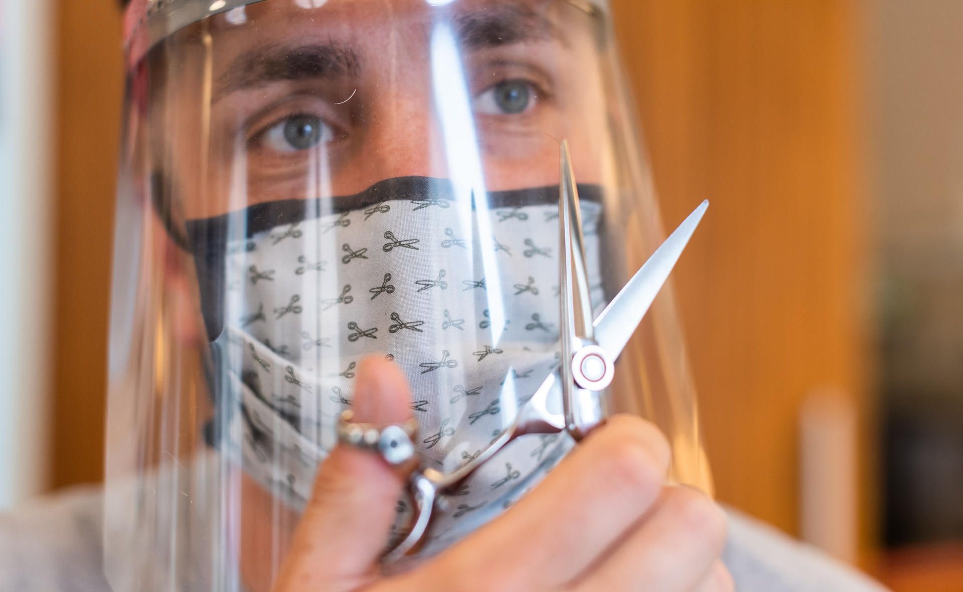 Dresden: Ein Friseur hält in einem Friseurstudio eine Haarschneideschere in der Hand und trägt dabei einen Mundschutz und ein Gesichtsschutzschild.
