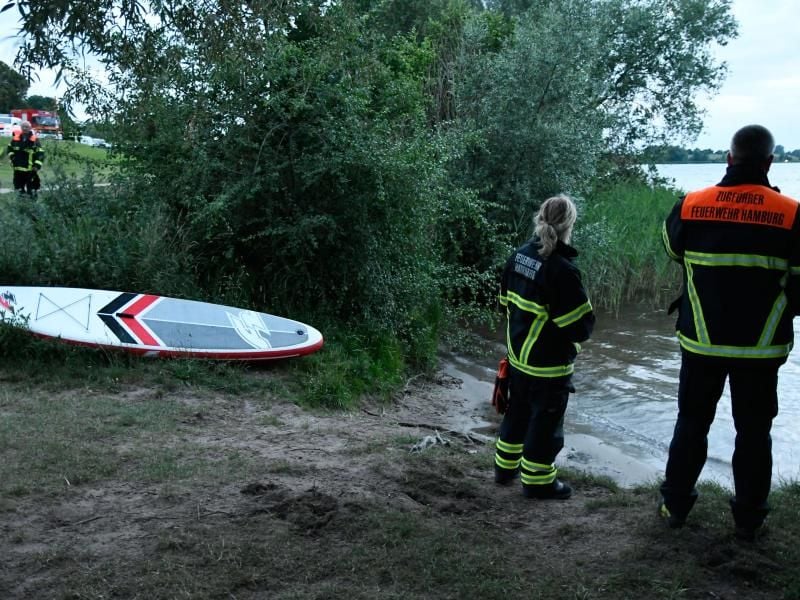 Einsatzkräfte der Feuerwehr blicken auf den Hohendeicher See, neben ihnen liegt ein Surfbrett. Foto: -/citynewstv
