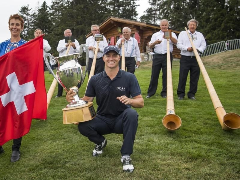 Der Schwede Alex Noren posiert mit seiner Trophäe zwischen Fahne mit Schweizerkreuz und Alphörnern. Foto: Jean-Christophe Bott