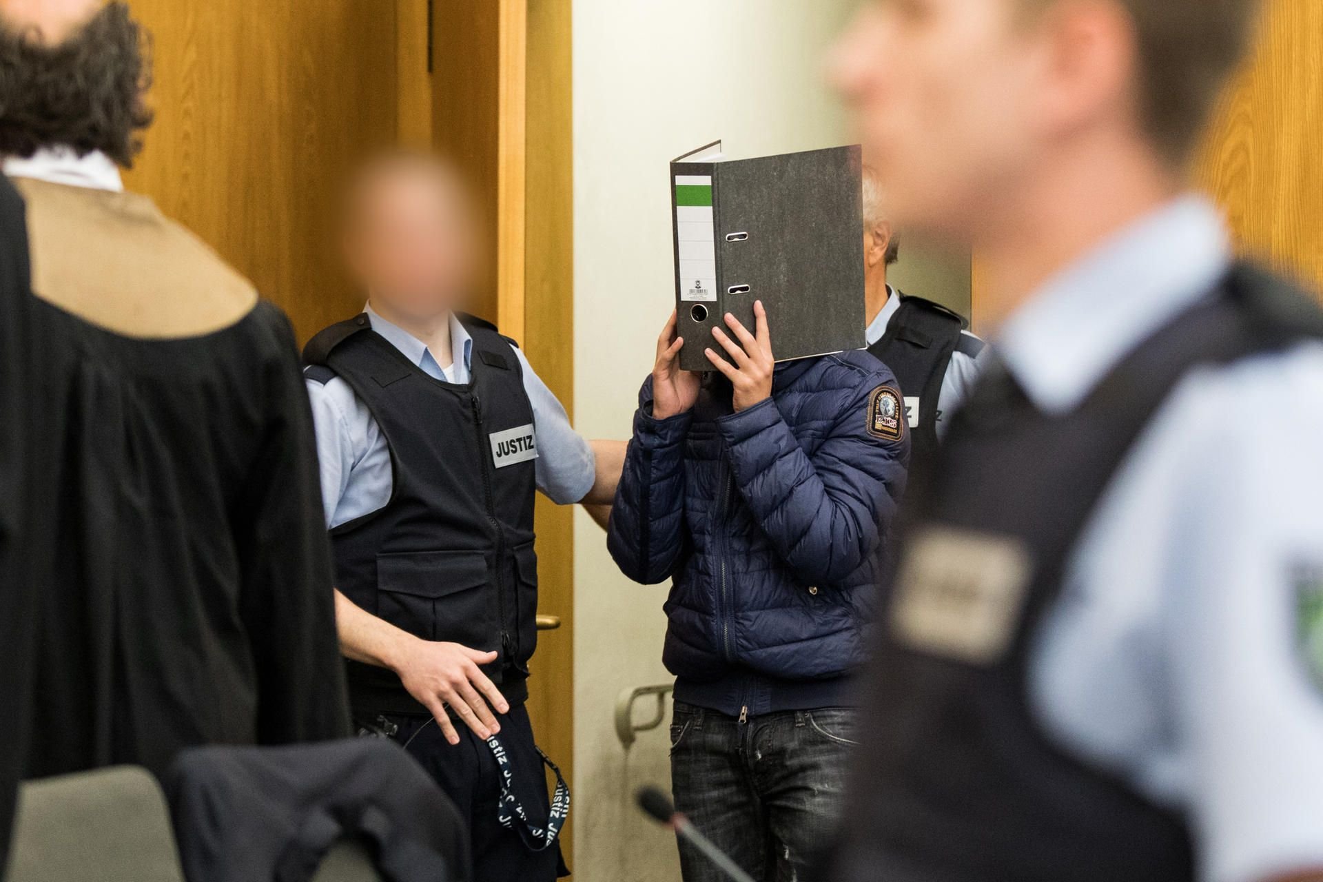 Einer der Angeklagten kommt mit einem Aktenordner vor dem Gesicht in den Gerichtssaal.