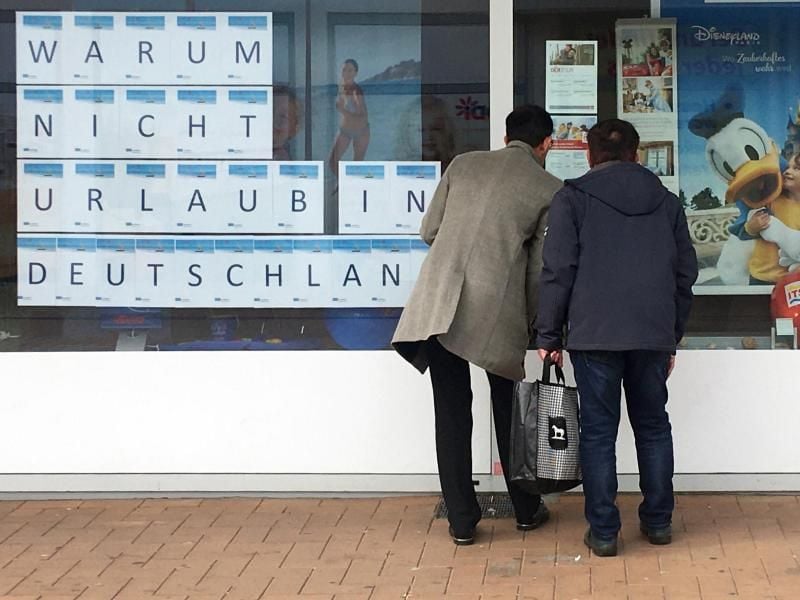 Zwei Männer schauen in das Schaufenster eines Reisebüros, an dem mit großen Buchstaben „Warum nicht Urlaub in Deutschland” zu lesen ist. Foto: Bernd Thissen/dpa