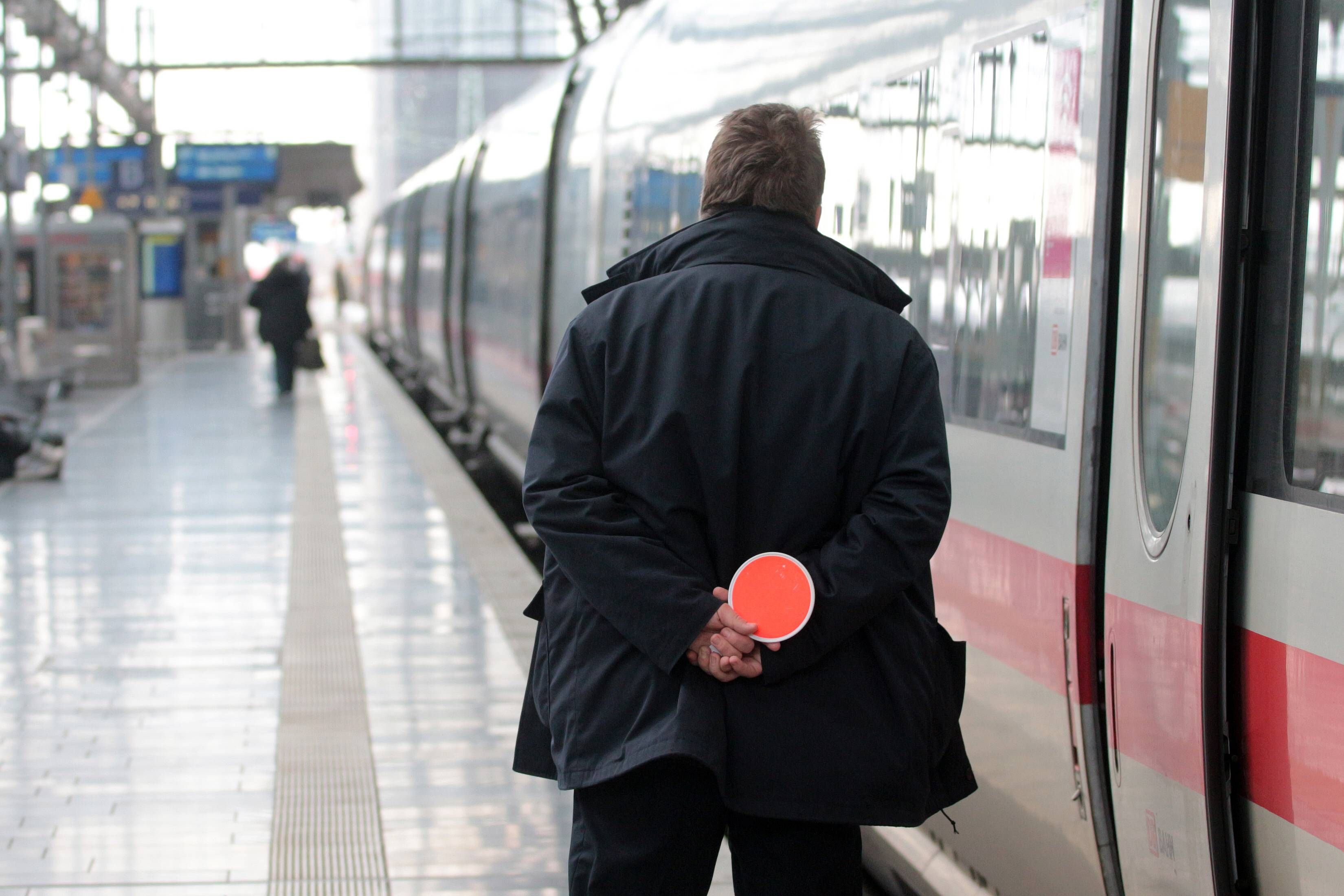 Ein Schaffner steht mit roter Kelle vor einem Zug am Bahnsteig.