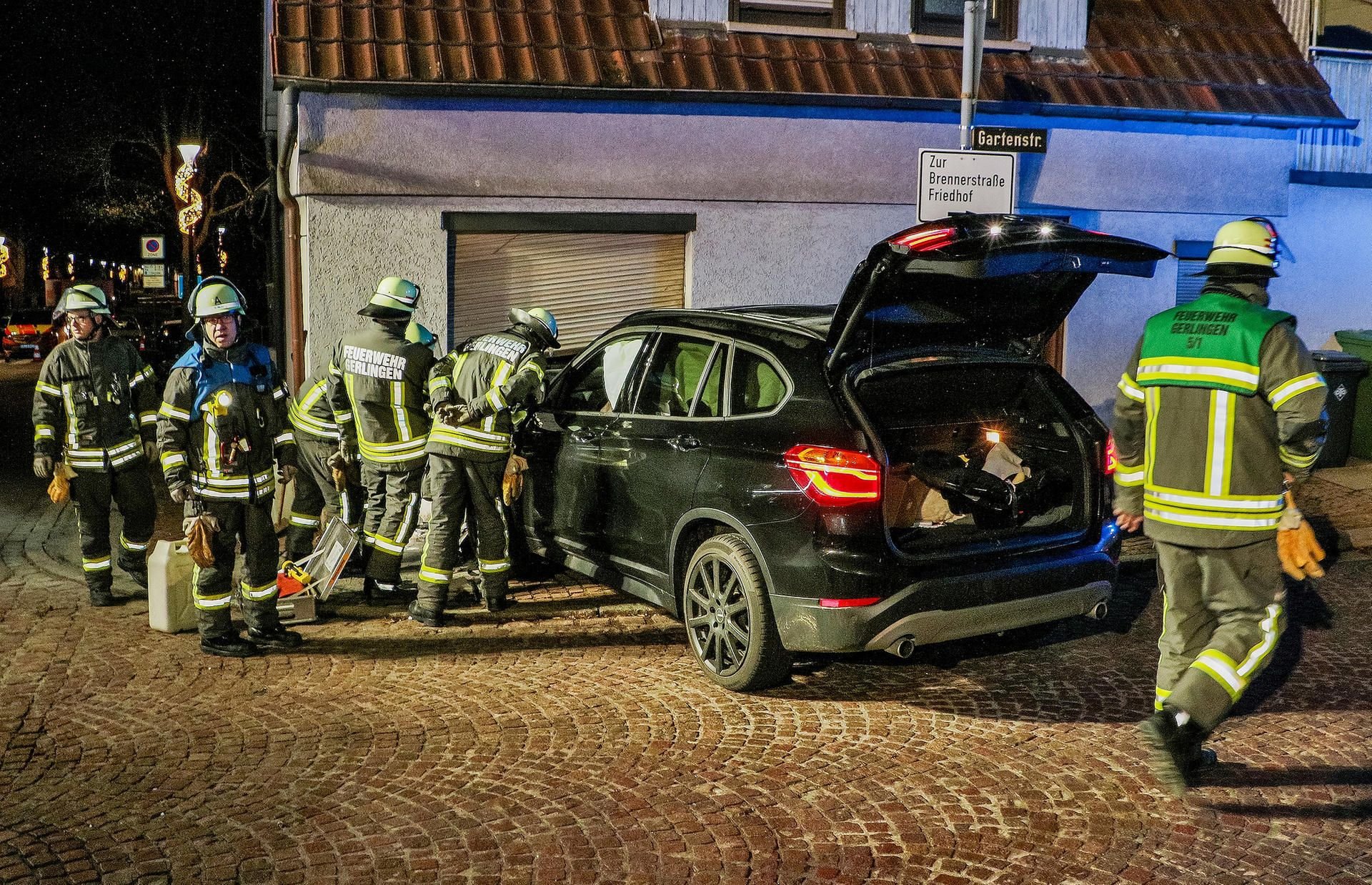 Feuerwehrleute stehen neben einem Auto mit zerstörter Motorhaube vor einer stellenweise zertrümmerten Wohnzimmerwand eines Einfamilienhauses in Gerlingen.