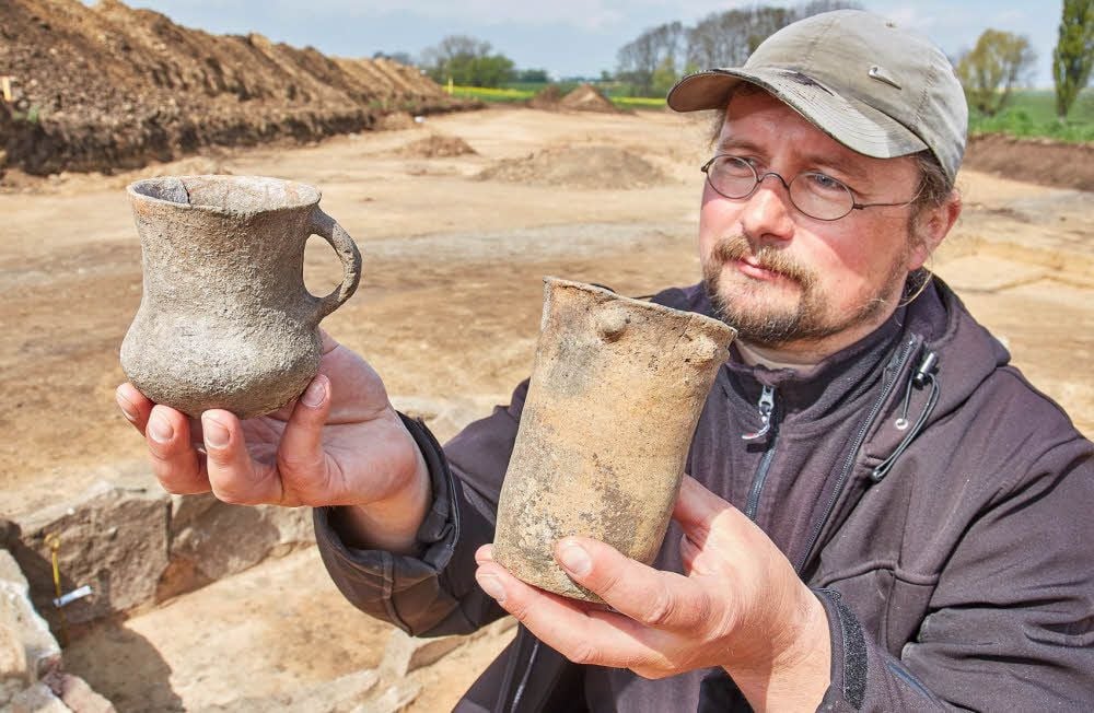 Grabungsleiter Thomas Kubenz zeigt vor einem der entdeckten Steingräber zwei Becher, die ebenfalls gefunden wurden.
