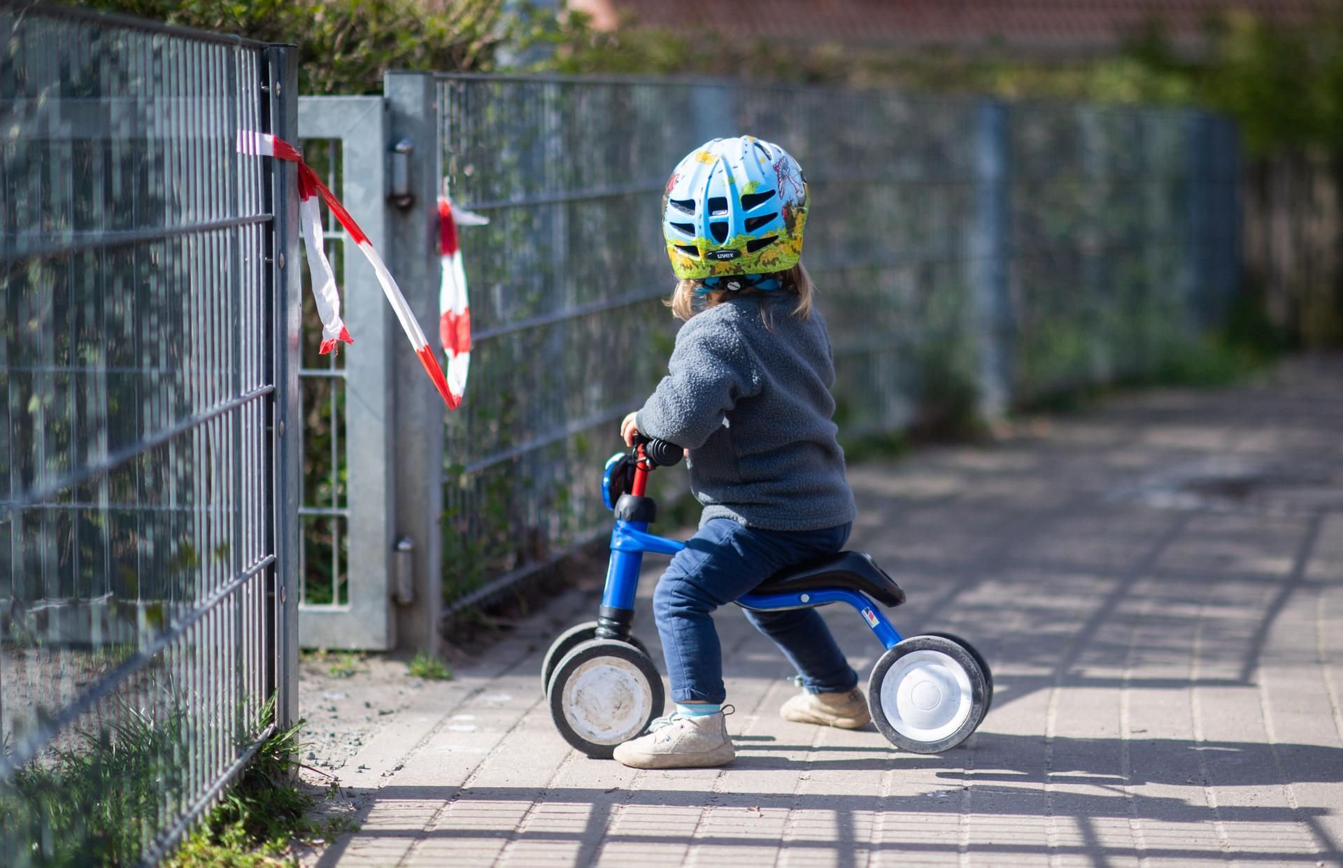 Ein Kleinkind steht mit einem Dreirad vor einem abgesperrten Spielplatz.