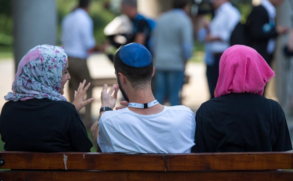Zwei junge Muslimas mit Kopftuch und ein junger jüdischer Mann mit Kippa unterhalten sich miteinander. (Symbolbild)