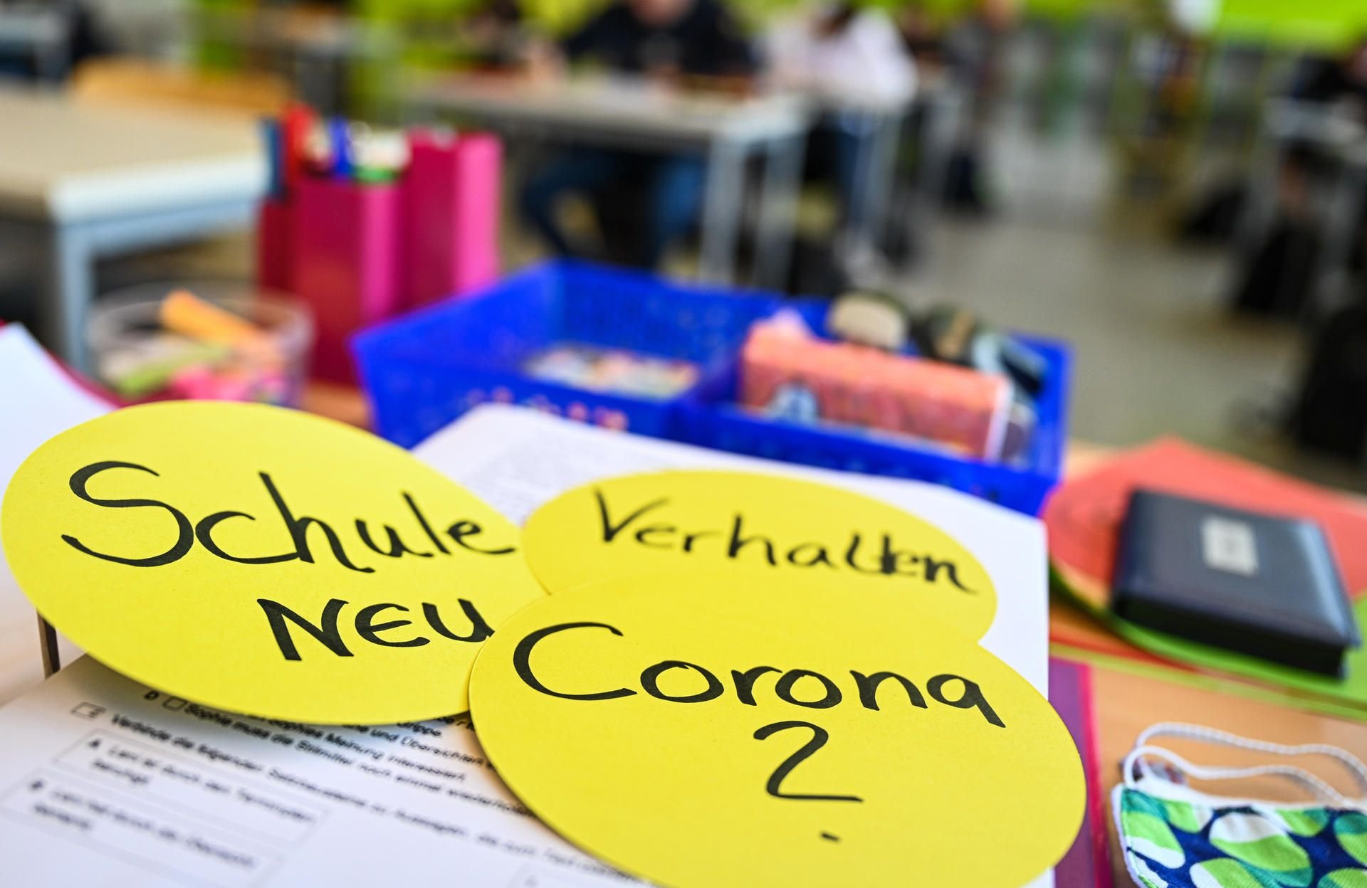 Schüler sitzen in einem Klassenzimmer, während auf dem Lehrerpult drei kreisrunde Papierschilder mit der Aufschrift Schule neu, Verhalten und Corona liegen.