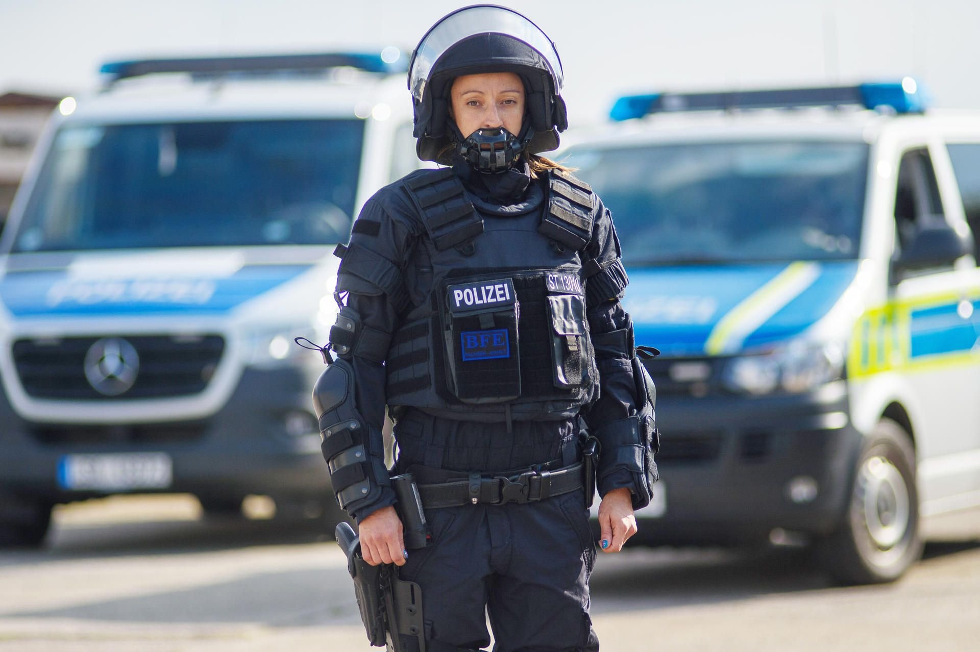 Eine Polizistin, ausgerüstet mit einer neuen Körperschutzausstattung (KSA), steht vor Polizeifahrzeugen.
