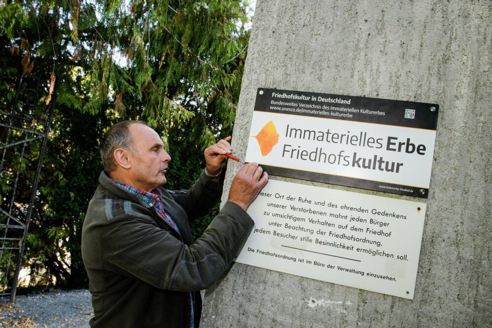 Friedhofsverwalter Andreas Legall befestigt am Eingangsbereich des Neuen Friedhofs ein Schild, das auf das Immaterielle Erbe Friedhofskultur hinweist. Auch Aushänge informieren darüber.