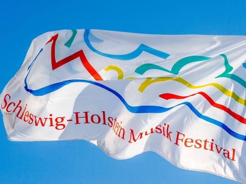 Eine Fahne mit dem Logo des Schleswig-Holstein Musik Festivals (SHMF) weht vor blauem Himmel. Foto: Markus Scholz/Archiv