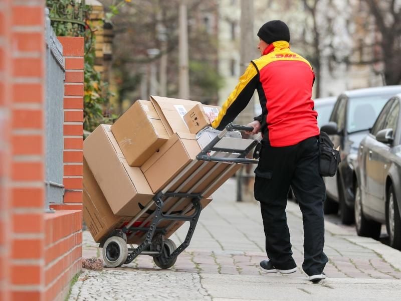 Ein DHL-Paketzusteller geht mit einer Sackkarre voller Pakete zu einem Haus. Foto: Jan Woitas/zb/dpa