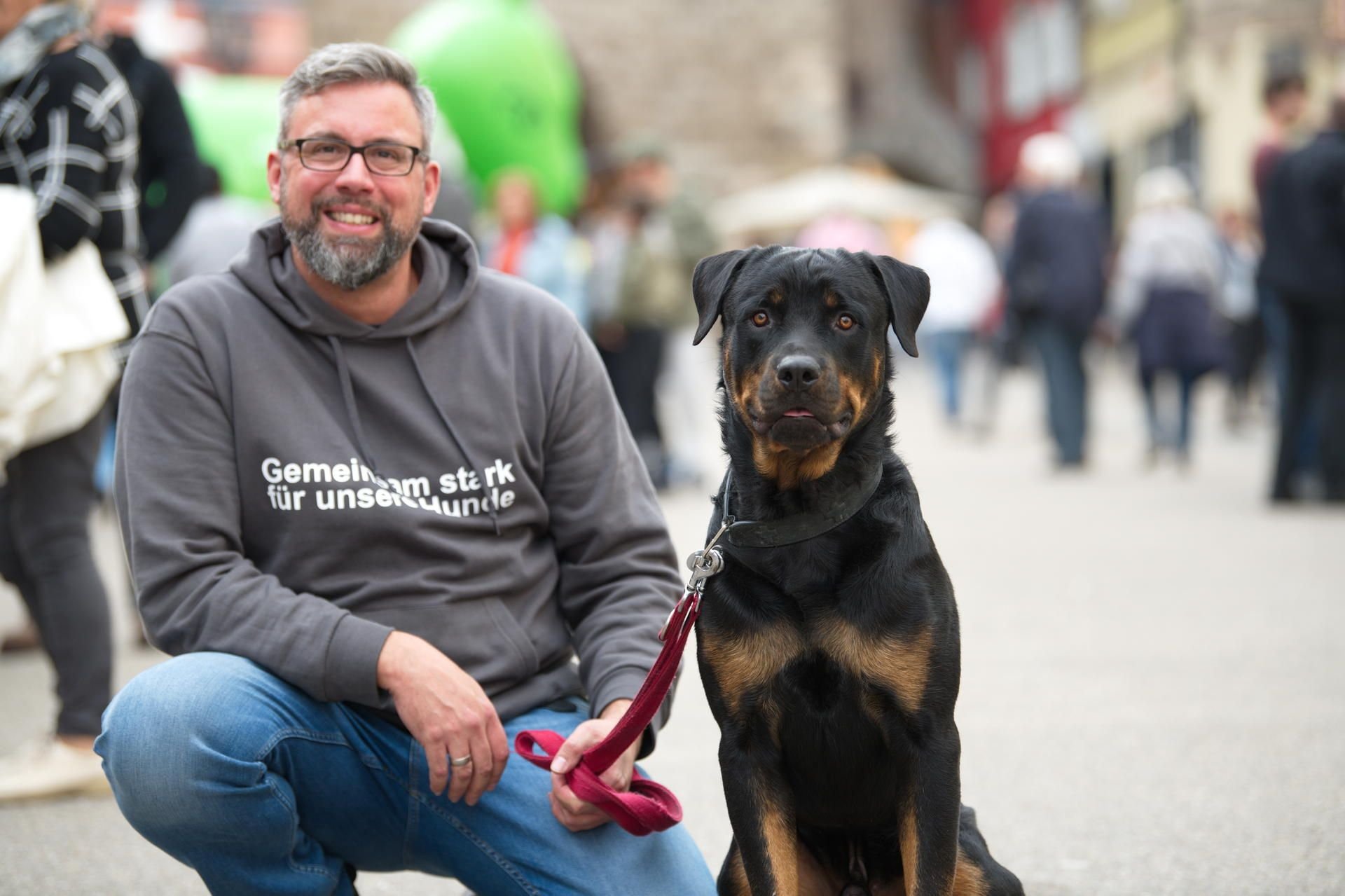 Markus hockt neben seinem Rottweiler während des Großen Rottweiler-Treffens unter dem Motto „Gemeinsam stark für unsere Hunde“, welches auch auf seinem Pullover geschrieben steht, in der Innenstadt von Rottweil.