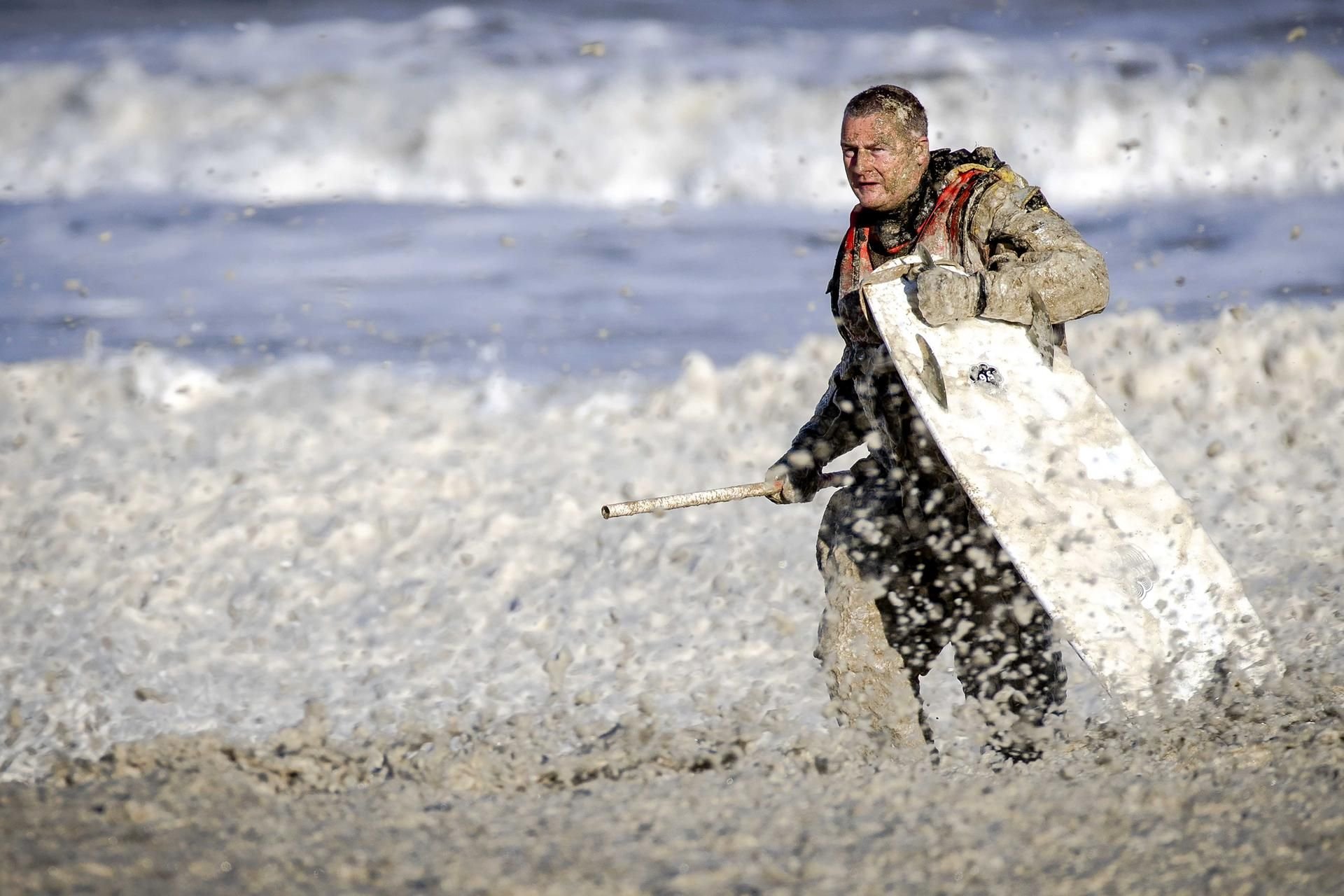 Ein Mitarbeiter der Seenotrettungsorganisation KNRM bedeckt von schmutzig-gelbem Schaum bergt ein Surfbrett.