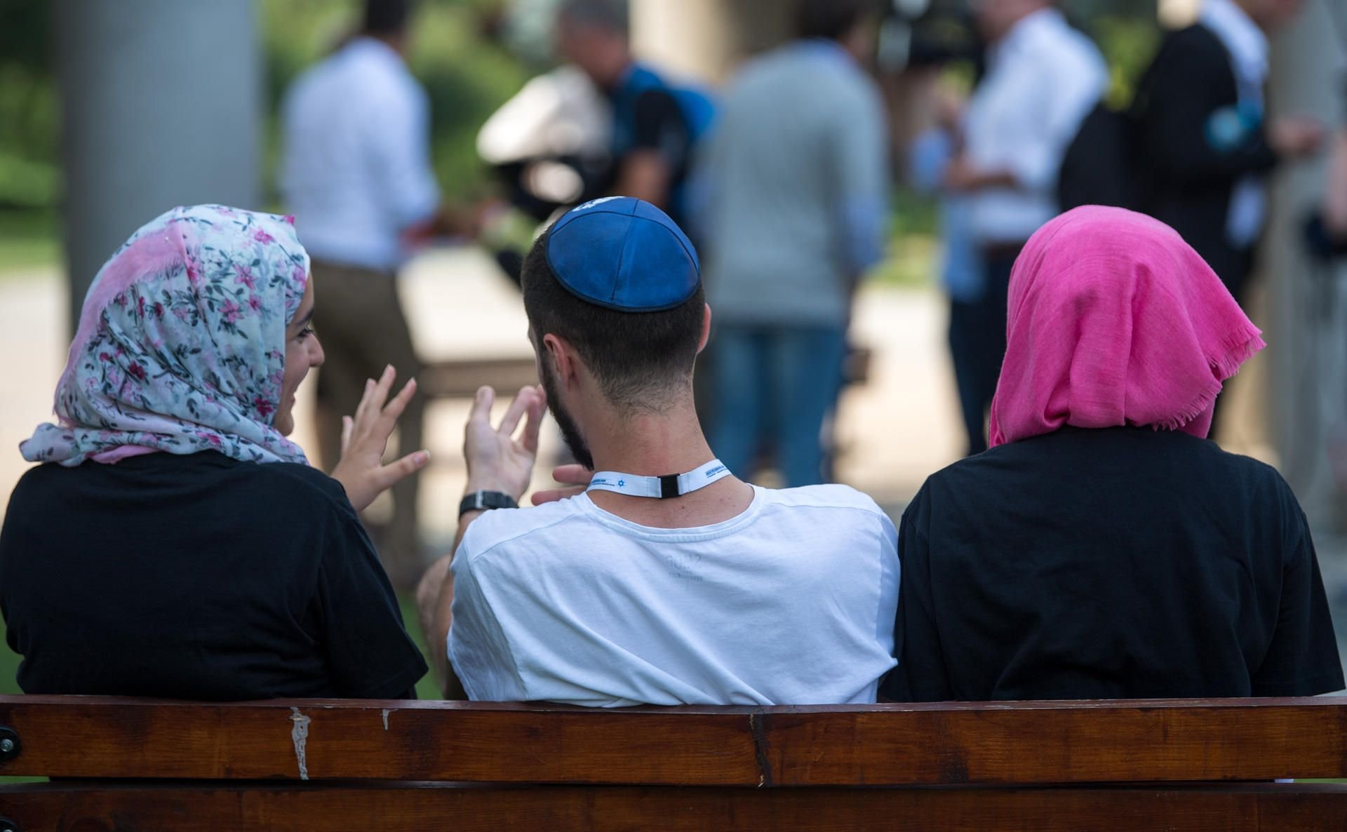 Zwei junge Muslimas mit Kopftuch und ein junger jüdischer Mann mit Kippa unterhalten sich miteinander. (Symbolbild)