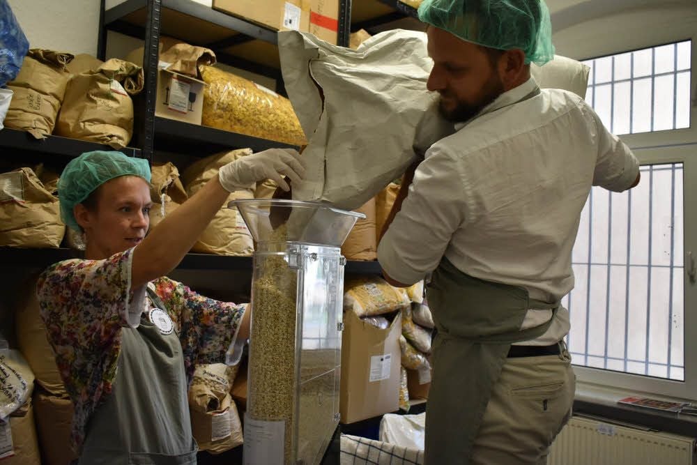 Jule Gottwald und Hannes Schulz füllen aus einem 25-Kilo-Sack aus Papier Haferflocken in ein Gefäß, das später im Verkaufsraum im Regal steht.