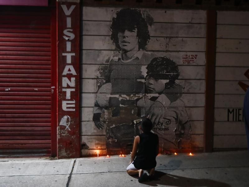 Ein Junge kniet vor einem mit Kerzen dekorierten Wandbild von Diego Maradona in Argentinien. Foto: Marcos Brindicci/AP/dpa