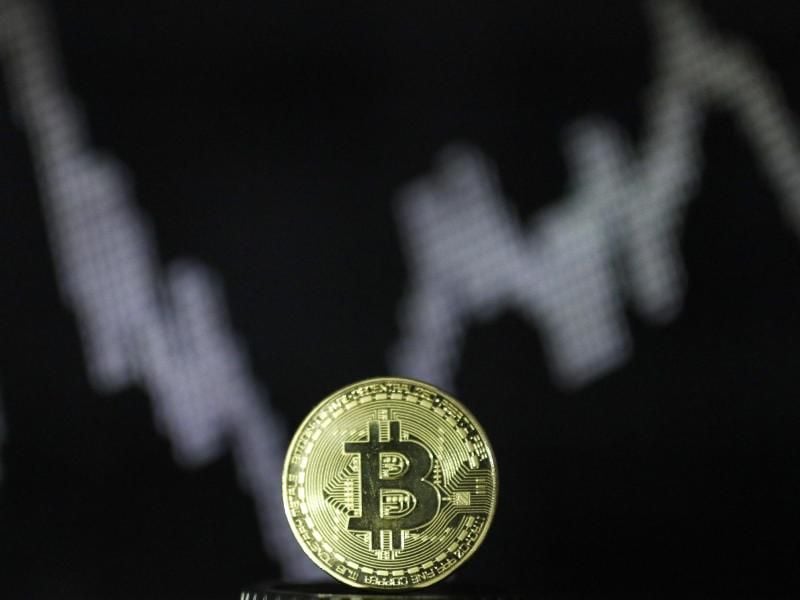 Eine Bitcoin-Münze ist vor einer Aktienkurve an einem Laptop zu sehen. Foto: Ina Fassbender/Archiv