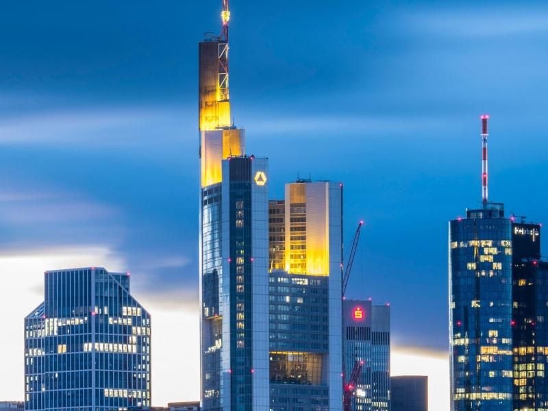 Wolken sind im Abendlicht hinter der Commerzbank (Mitte, mit gelber Beleuchtung) und anderen Hochhäusern der Bankenskyline zu sehen. Foto: Boris Roessler/dpa/Archivbild