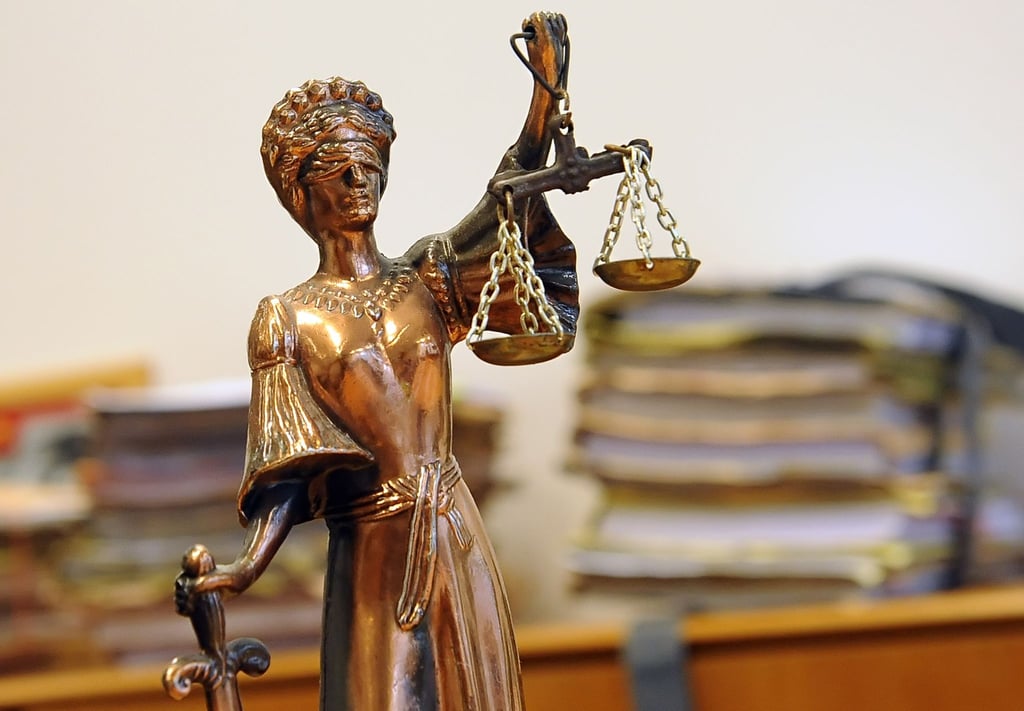 Eine goldfarbene Justitia-Figur steht vor Aktenbergen, die sich auf einem Tisch stapeln.
