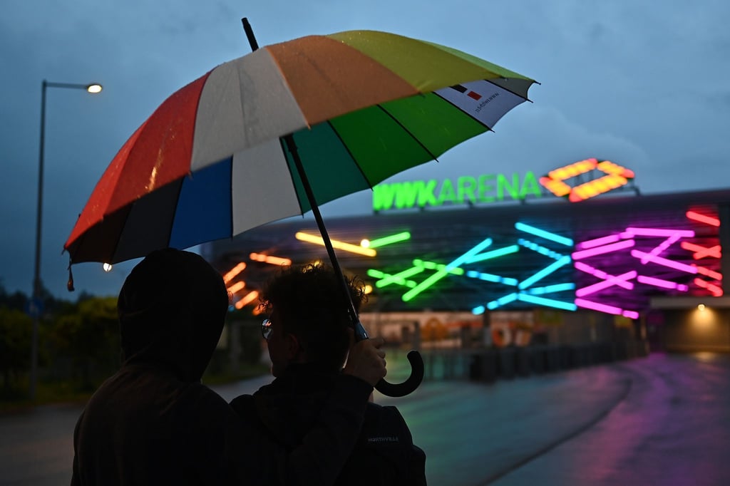 FußZwei junge Männer mit einem Regenbogen-Schirm stehen vor WWK-Arena in Augsburg, die während des Spiels in Regenbogenfarben beleuchtet ist.