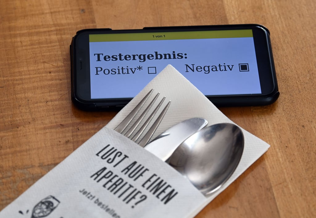 Ein Serviettenetui mit Aufdruck «Lust auf einen Aperitif?» liegt in einem Restaurant neben einem Smartphone, dessen Display ein negatives Corona-Testergebnis zeigt.