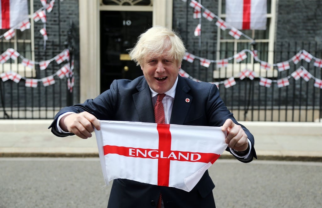 Premierminister Boris Johnson hält eine englische Fahne vor sich in die Höhe.