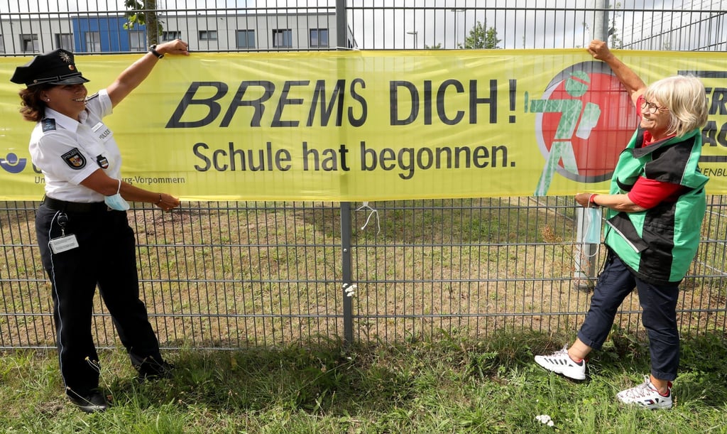Ein Banner mit der Aufschrift "Brems dich! Die Schule hat begonnen." wird an einem Zaun aufgehangen.