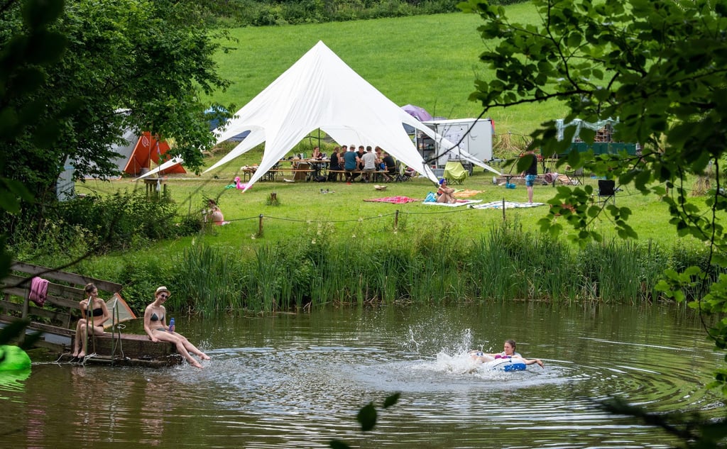 Teilnehmer eines Ferienlagers schwimmen in einem See, während die anderen zusammen im Gemeinschaftszelt sitzen.