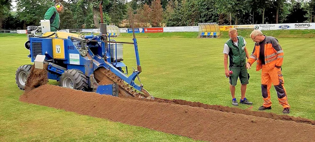 Bauarbeiter einer Spezialfirma ziehen auf dem Sportplatz in Velsdorf Gräben, in denen die Rohre für die Unterflurberegnungsanlage gelegt werden. Im September soll der Platz wieder bespielbar sein.