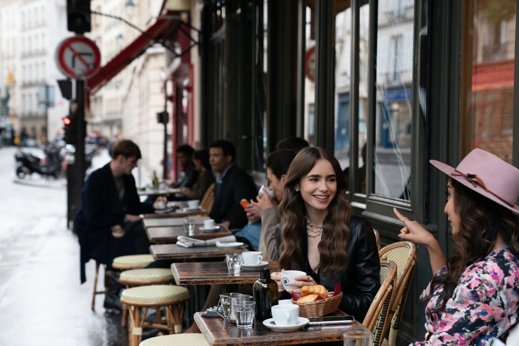 Emily (Lily Collins, m) sitzt mit einer Freundin im Café - Filmszene aus der ersten Staffel der Netflix-Serie „Emily in Paris“.