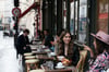 Emily (Lily Collins, m) sitzt mit einer Freundin im Café - Filmszene aus der ersten Staffel der Netflix-Serie „Emily in Paris“.