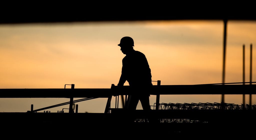 Die Silhouette eines Bauarbeiters zeichnet sich auf einer Baustelle vor dem verfärbten Morgenhimmel ab.