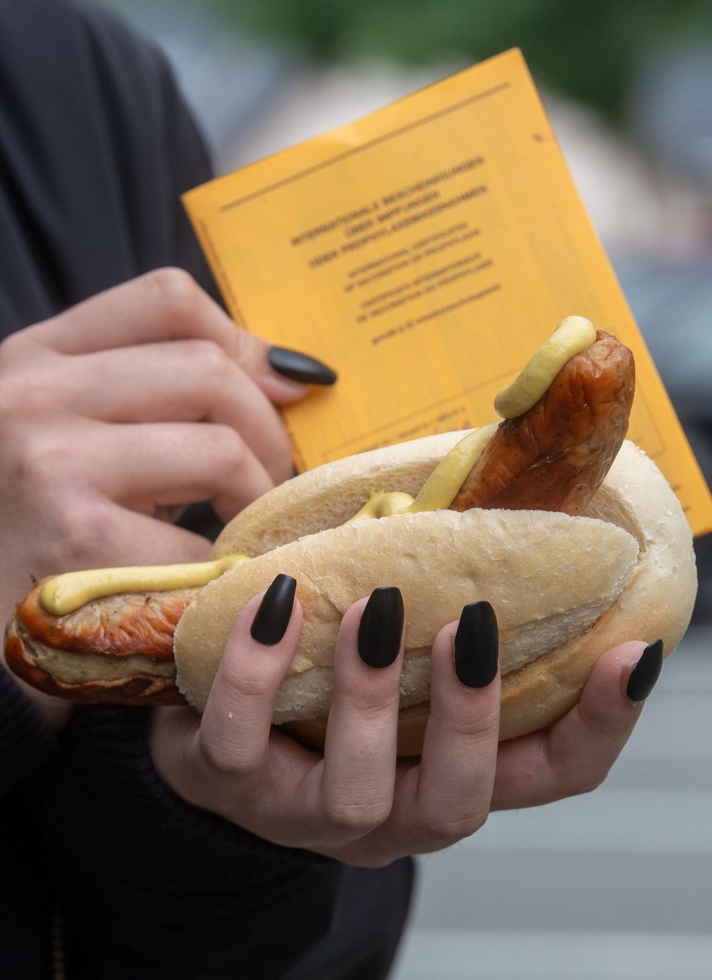 Eine junge Frau hält am mobilen Impfzentrum einen Impfpass und eine Bratwurst im Bötchen.