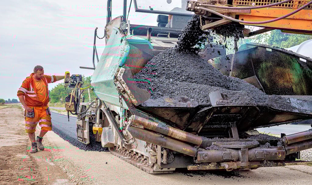 Knapp 160 Grad Celsius beträgt die Temperatur des Asphalts, wenn er aufgetragen wird. Hier überwacht René Voß, dass die acht Zentimeter starke und 2,3 Meter breite Tragschicht korrekt vom Asphaltfertiger - so heißt diese Straßenbaumaschine - aufgebracht wird.