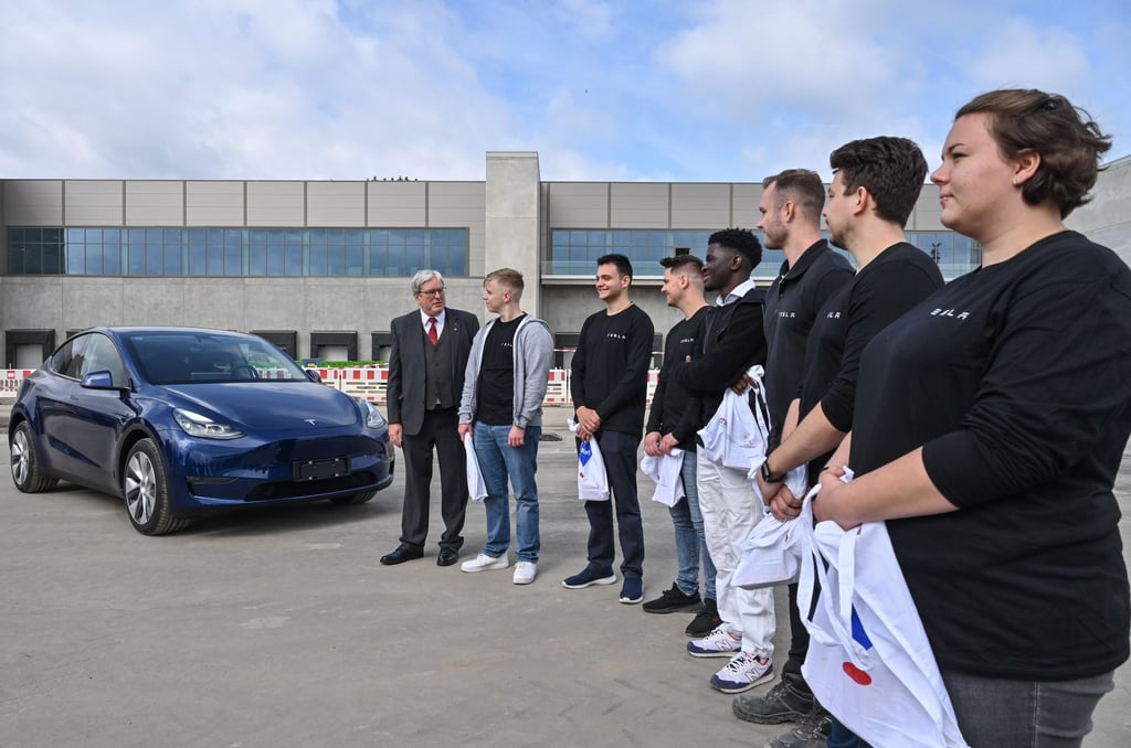 Wirtschaftsminister Jörg Steinbach (SPD, l) steht neben einem Tesla und den ersten Auszubildenden.