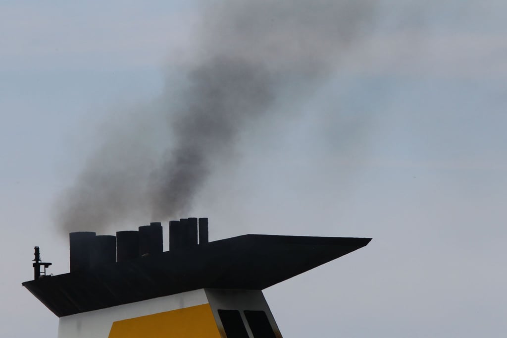 Eine dunkle Abgaswolke steigt aus dem Schornstein eines Fährschiffs bei der Ausfahrt aus dem Seehafen.